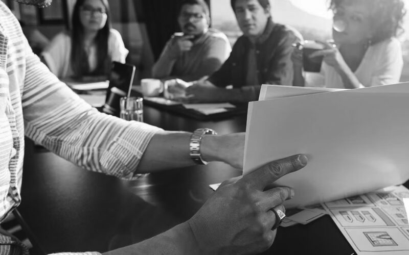 A person reviews documents at a conference table while four colleagues sit across, discussing the Perfect Fit for a new IT Model. Papers and glasses of water are on the table, and sunlight streams through a window.