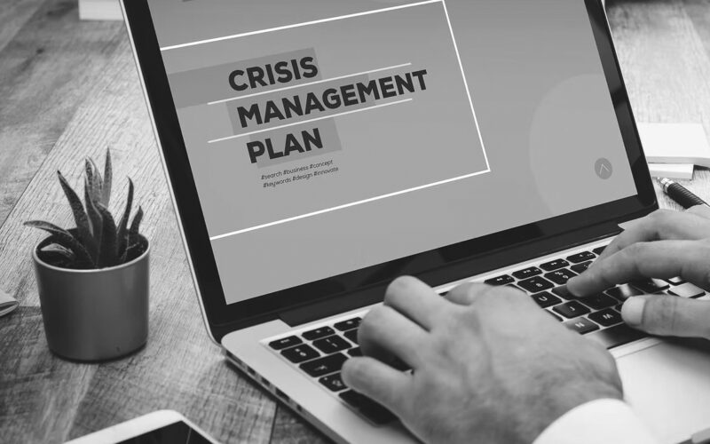 A person types on a laptop displaying a screen with the words Crisis Management Plan. A small potted plant, a notebook, and papers are visible on the wooden desk, highlighting the importance of disaster recovery planning for bonus benefits.