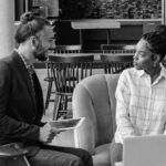 Two people sit indoors having a conversation; one holds a tablet and smiles, while the other sits next to a laptop, listening attentively. They appear to be in a casual office or coworking space focused on business productivity.