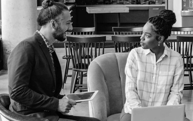 Two people sit indoors having a conversation; one holds a tablet and smiles, while the other sits next to a laptop, listening attentively. They appear to be in a casual office or coworking space focused on business productivity.