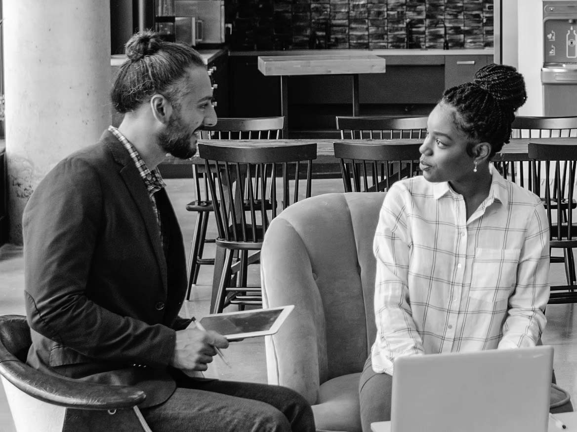 Two people sit indoors having a conversation; one holds a tablet and smiles, while the other sits next to a laptop, listening attentively. They appear to be in a casual office or coworking space focused on business productivity.
