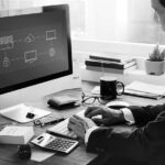 A man in a suit works at a desk with a large computer monitor displaying a business network diagram, planning for backup and disaster recovery. The desk has office supplies, a calculator, cup, notebooks, and a small potted plant.