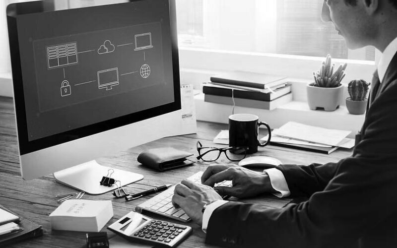A man in a suit works at a desk with a large computer monitor displaying a business network diagram, planning for backup and disaster recovery. The desk has office supplies, a calculator, cup, notebooks, and a small potted plant.