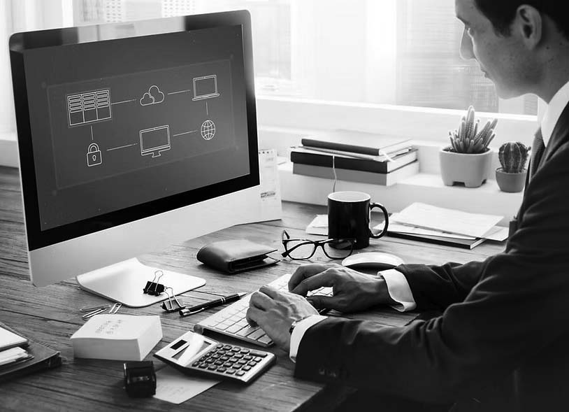 A man in a suit works at a desk with a large computer monitor displaying a business network diagram, planning for backup and disaster recovery. The desk has office supplies, a calculator, cup, notebooks, and a small potted plant.