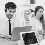 Two customer service representatives wearing headsets sit at a desk, working on laptops for emergency IT help desk support. Another person in the foreground holds a tablet in the bright, modern office environment.