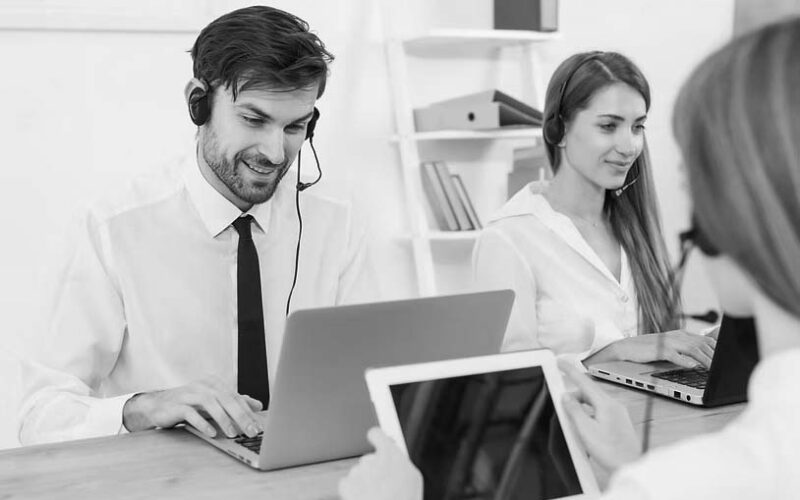 Two customer service representatives wearing headsets sit at a desk, working on laptops for emergency IT help desk support. Another person in the foreground holds a tablet in the bright, modern office environment.