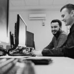 Two young men sit side by side at a desk in a modern, well-lit office, smiling and working together on desktop computers—demonstrating reliable IT support from Pavelcomm. Multiple monitors highlight their efficient business IT support setup.