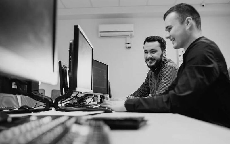 Two young men sit side by side at a desk in a modern, well-lit office, smiling and working together on desktop computers—demonstrating reliable IT support from Pavelcomm. Multiple monitors highlight their efficient business IT support setup.