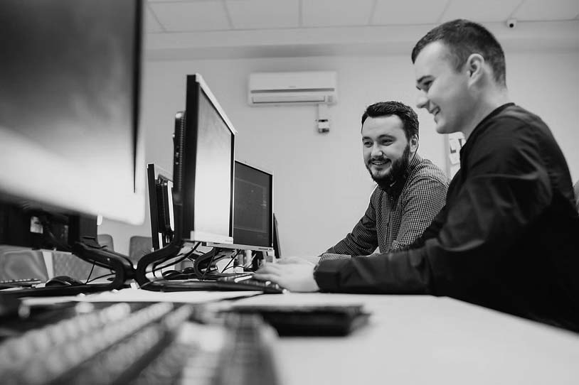 Two young men sit side by side at a desk in a modern, well-lit office, smiling and working together on desktop computers—demonstrating reliable IT support from Pavelcomm. Multiple monitors highlight their efficient business IT support setup.