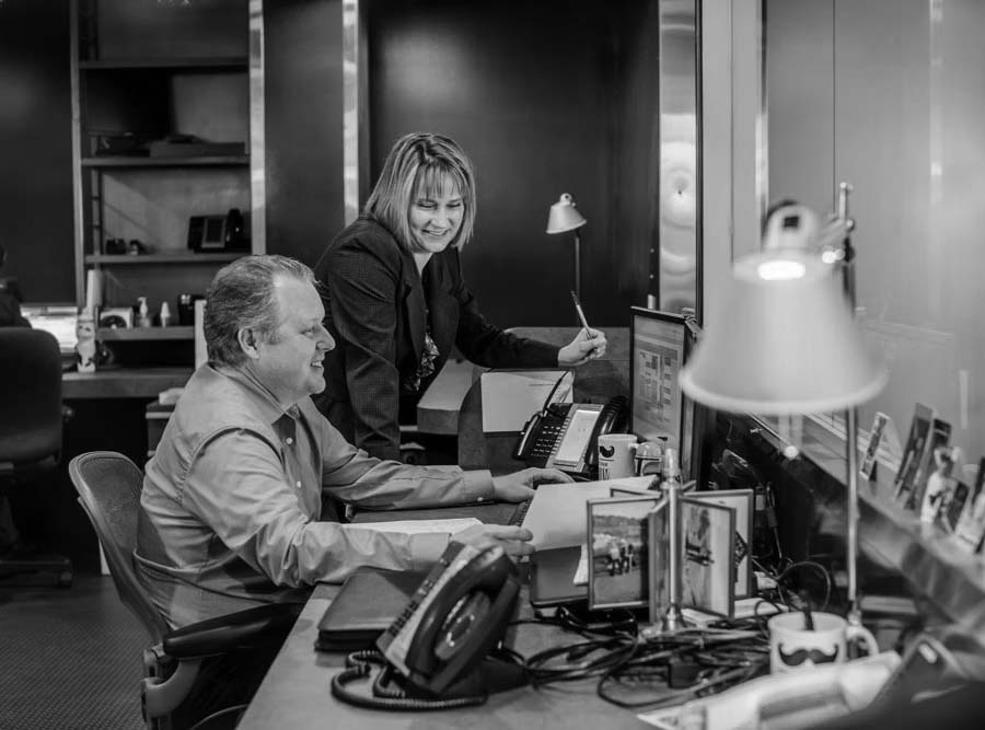 Two office workers, a man sitting and a woman standing, smile as they look at computer screens in a modern, red-walled office, showcasing Pavelcomm’s exceptional IT Services surrounded by phones, papers, and lamps.