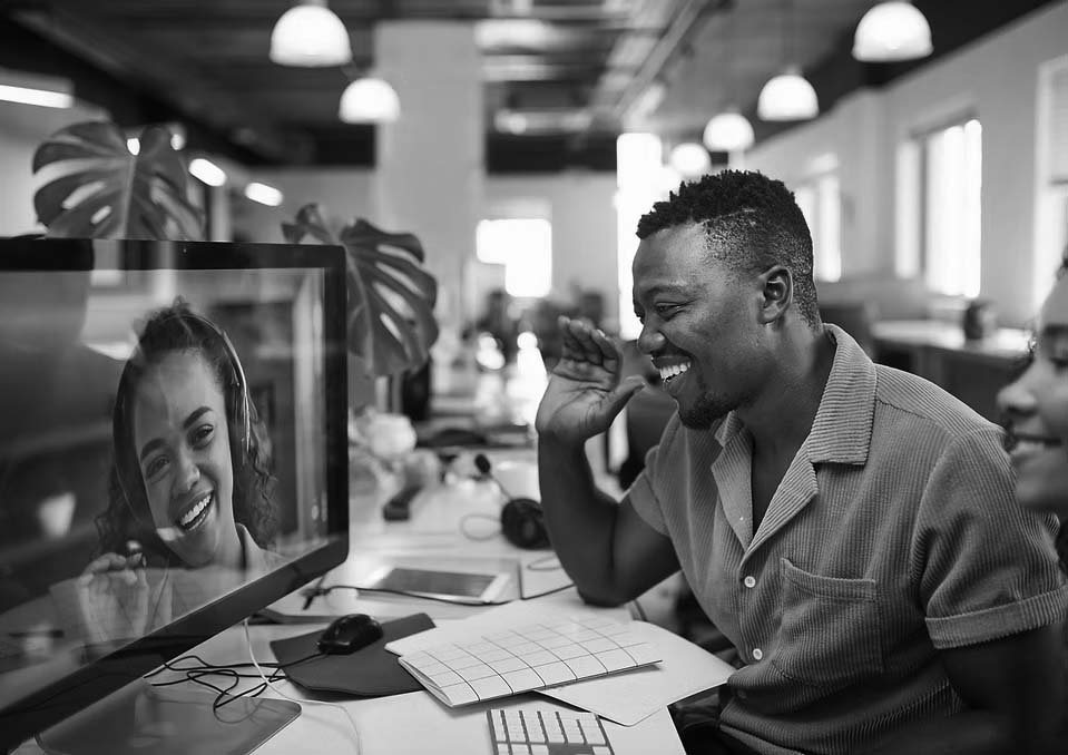 A man in a yellow shirt sits at a desk, smiling and waving at a woman on a video call displayed on his computer monitor in a modern office setting.
