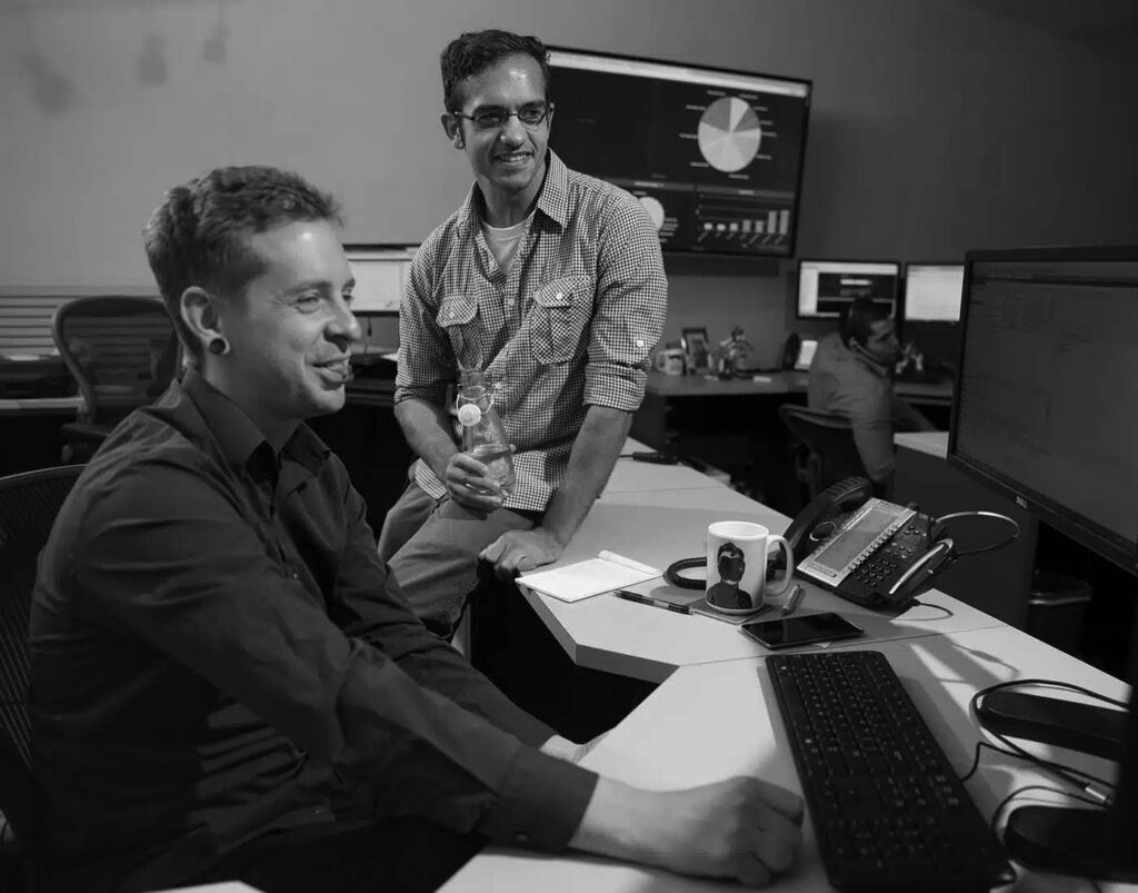 pavelcomm-helpdesk-new-1.jpg Two men working at computer desks in an office, one sitting and using the keyboard, the other standing and smiling. Large monitors display charts and data, highlighting a busy Emergency Help Desk environment.