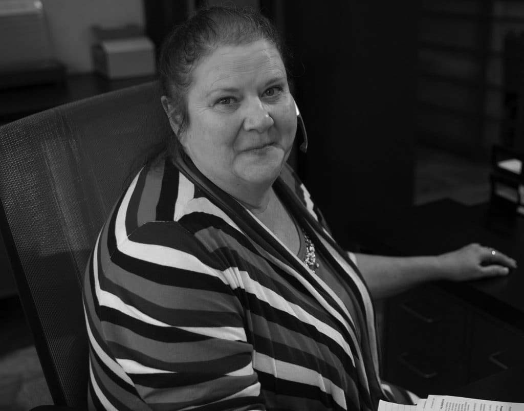 A woman with light skin and brown hair, wearing a blue, teal, and white striped top, sits at an office desk with papers and a headset, smiling at the camera—demonstrating a professional setting focused on HIPAA compliance.