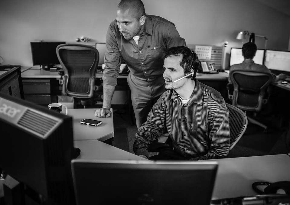 Two men in an office are focused on a computer screen; one is sitting with a headset, while the other stands beside him, leaning in to assist. Other employees work at computers in the background at Pavelcomm, specialists in IT Services.