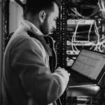 A man in a yellow jacket works on a laptop while standing next to a server rack filled with cables and equipment in a data center.