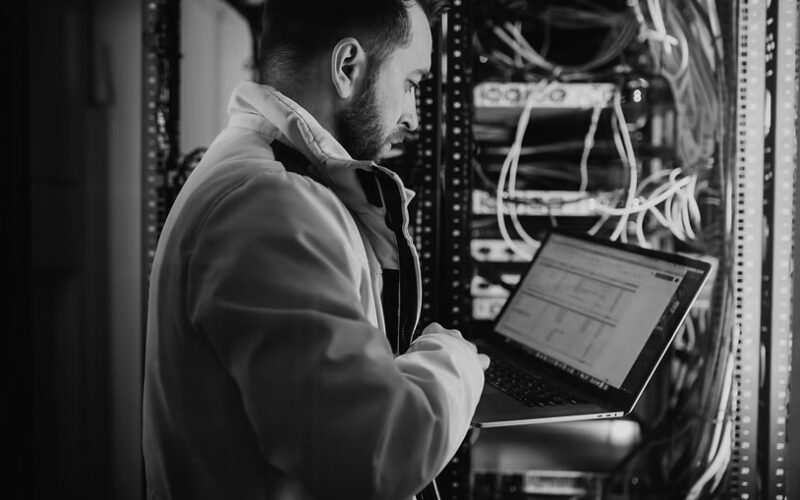 A man in a yellow jacket works on a laptop while standing next to a server rack filled with cables and equipment in a data center.