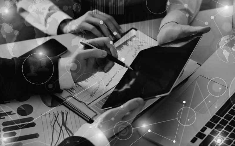 Three people collaborate at a desk with charts, a laptop, and a tablet, discussing data and analytics. Digital graphics highlight cloud computing and network connections, emphasizing teamwork and the advantages of technology in business analysis.