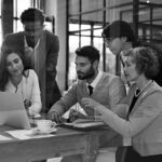 Five colleagues gather around a wooden table in a modern office, discussing work with a laptop open to IT services. Papers, a notebook, and a pen are on the table. The atmosphere appears collaborative and professional.