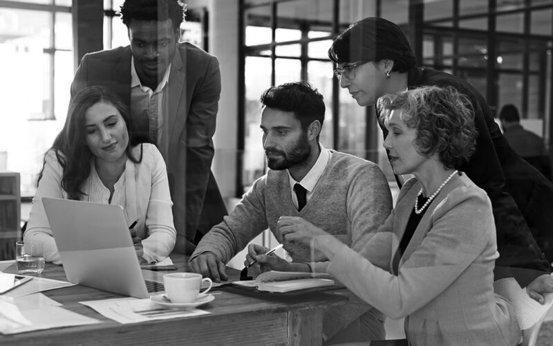 Five colleagues gather around a wooden table in a modern office, discussing work with a laptop open to IT services. Papers, a notebook, and a pen are on the table. The atmosphere appears collaborative and professional.