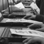 Rows of people in a Portland classroom work on laptops with printed documents, actively typing and using mice. Papers with text and diagrams suggest a hands-on cybersecurity training session to integrate cybersecurity skills.