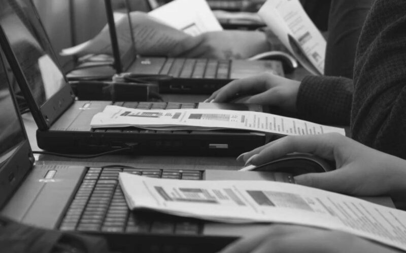 Rows of people in a Portland classroom work on laptops with printed documents, actively typing and using mice. Papers with text and diagrams suggest a hands-on cybersecurity training session to integrate cybersecurity skills.