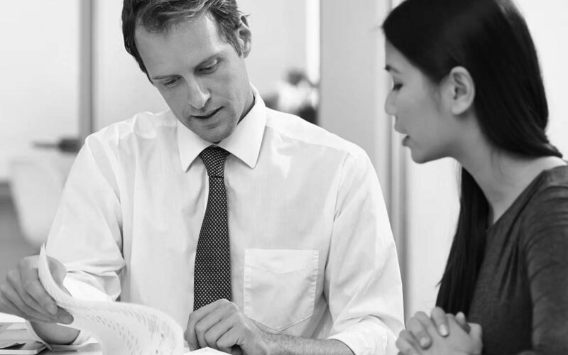 A man in a white shirt and tie shows papers to a woman with long dark hair. They are sitting at a table in an office, looking focused while discussing cloud compliance solutions for their business.