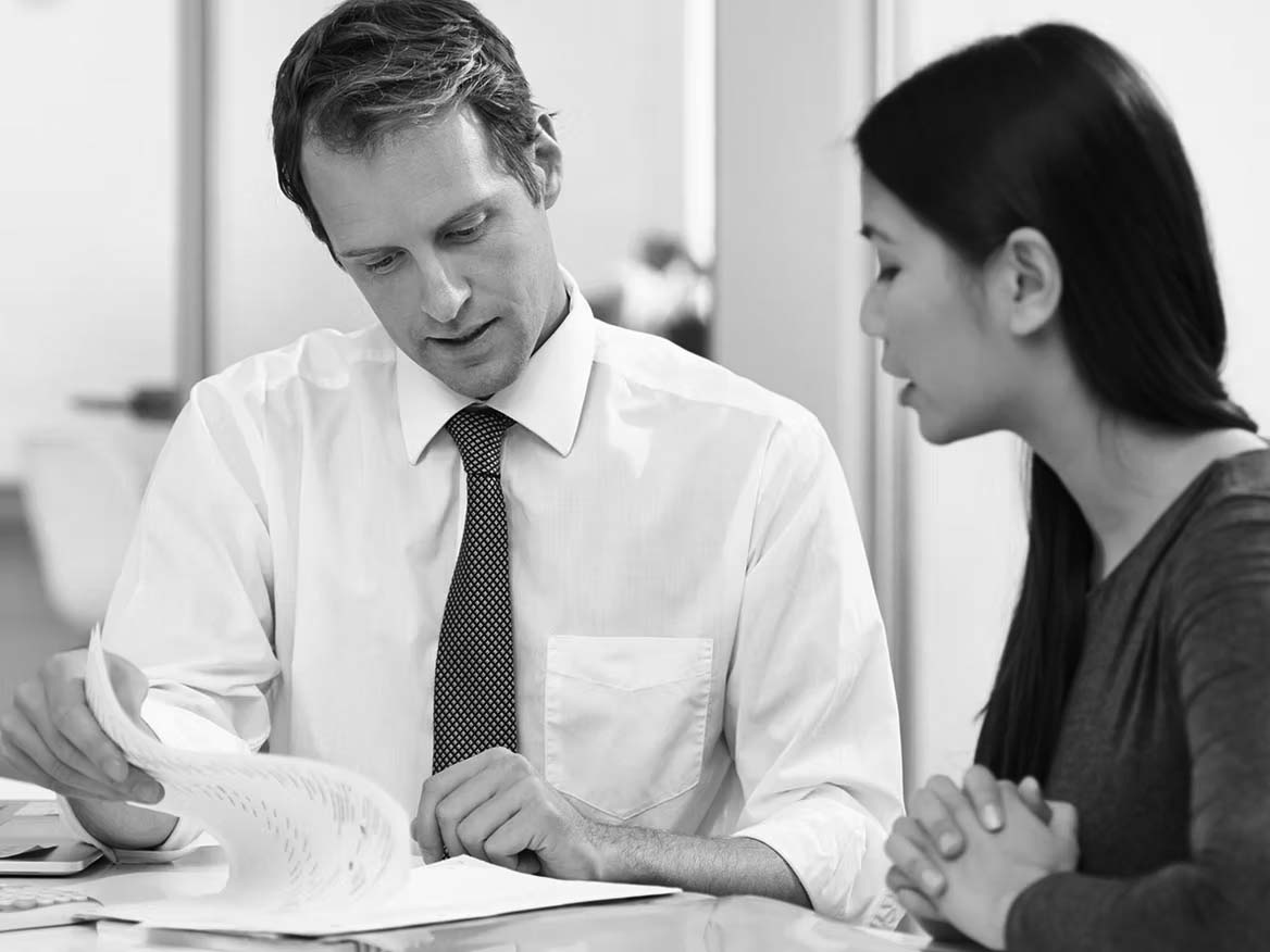 A man in a white shirt and tie shows papers to a woman with long dark hair. They are sitting at a table in an office, looking focused while discussing cloud compliance solutions for their business.