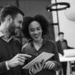 Two coworkers, a man and a woman, smiling and looking at a tablet together in a modern office—perhaps exploring the benefits of cloud communications. In the background, two other colleagues work at a desk with laptops.