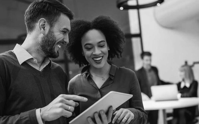 Two coworkers, a man and a woman, smiling and looking at a tablet together in a modern office—perhaps exploring the benefits of cloud communications. In the background, two other colleagues work at a desk with laptops.