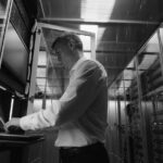 A man in a white shirt stands in a server room, working on a laptop mounted in a server rack. Surrounded by blinking lights, he focuses on cyber security to help the data center regain control after a potential cyberattack.