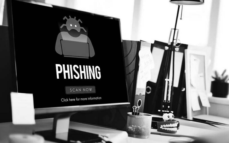 A computer screen displays an IT security phishing warning with a cartoon robot and the words PHISHING plus a red SCAN NOW button. The desk has office supplies, a coffee cup, and a red toy with teeth to help prevent cyber attacks.