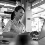 A woman sits at a table in a bright café, smiling as she works on cloud computing from her laptop. A cup of coffee is on the table in front of her, and large windows and a blurred person standing are seen in the background.
