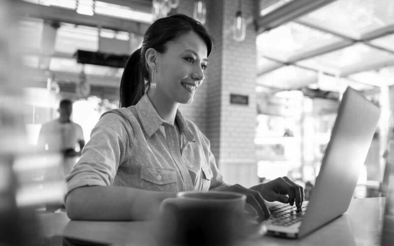 A woman sits at a table in a bright café, smiling as she works on cloud computing from her laptop. A cup of coffee is on the table in front of her, and large windows and a blurred person standing are seen in the background.