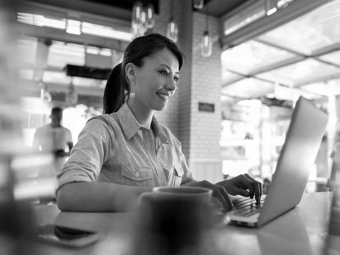 A woman sits at a table in a bright café, smiling as she works on cloud computing from her laptop. A cup of coffee is on the table in front of her, and large windows and a blurred person standing are seen in the background.