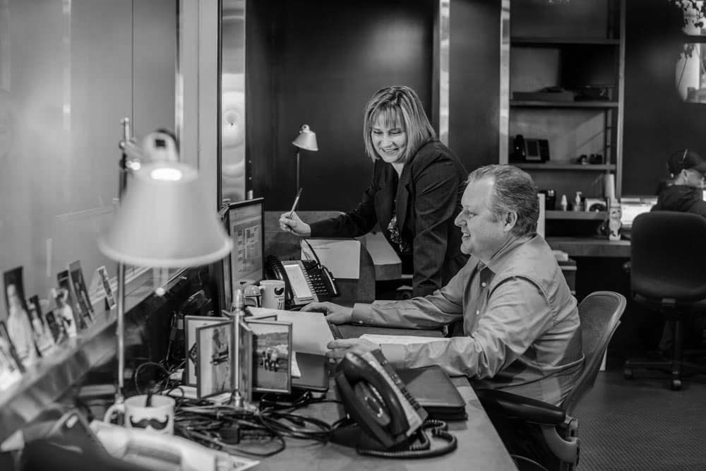 Two people in business attire work at a desk with computers in a modern office, collaborating on customer solutions. One types while the other stands, smiling and holding a pen. Office equipment and framed photos are visible on the desk.