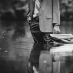 A man in a tan colored suit, in a nature setting, standing in a large puddle of water, holding a bag meant to hold files, as it gently floats on top of the water.