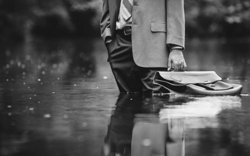 A man in a tan colored suit, in a nature setting, standing in a large puddle of water, holding a bag meant to hold files, as it gently floats on top of the water.