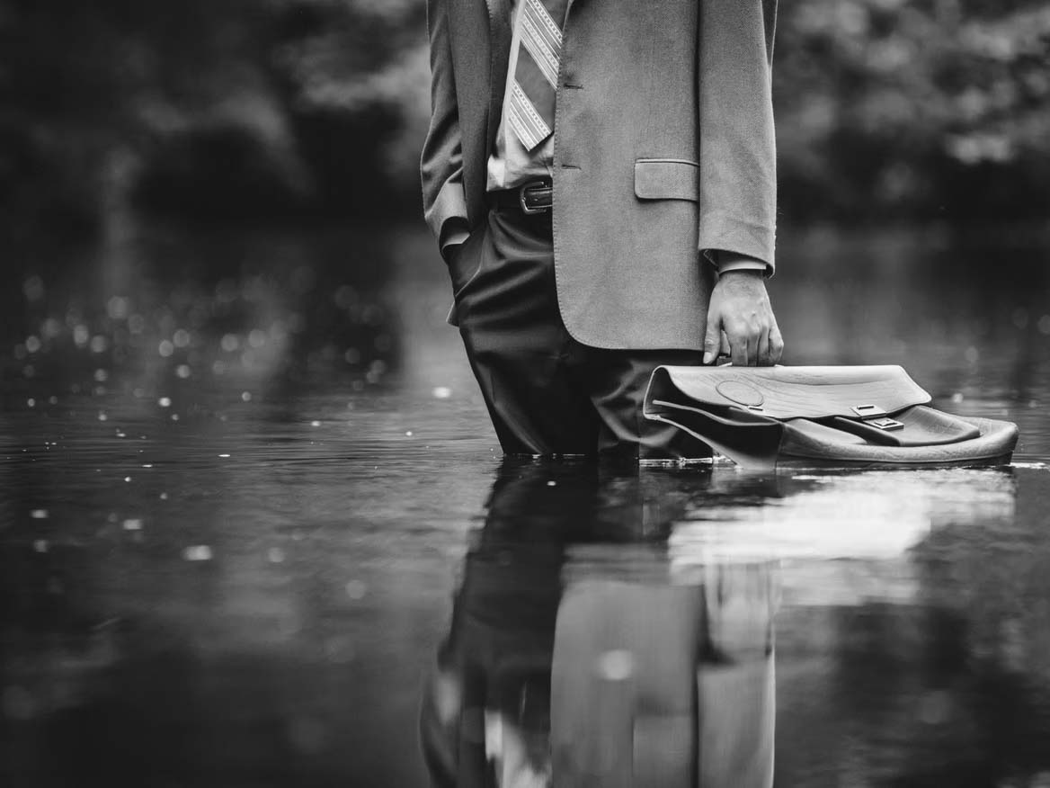 A man in a tan colored suit, in a nature setting, standing in a large puddle of water, holding a bag meant to hold files, as it gently floats on top of the water.