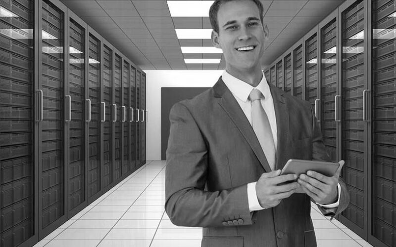 A smiling man in suit standing in a server room, looking into the camera while holding a tablet.
