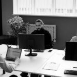 An office setting with several people sitting around laptop and desktop computers thinking and looking at their workstations.