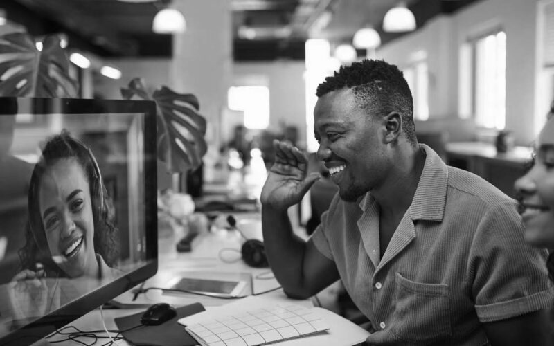A man in an office setting speaking and laughing with another person being transmitted via networking devices and monitor.