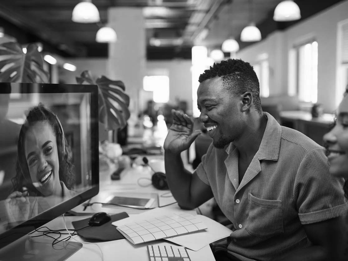 A man in an office setting speaking and laughing with another person being transmitted via networking devices and monitor.