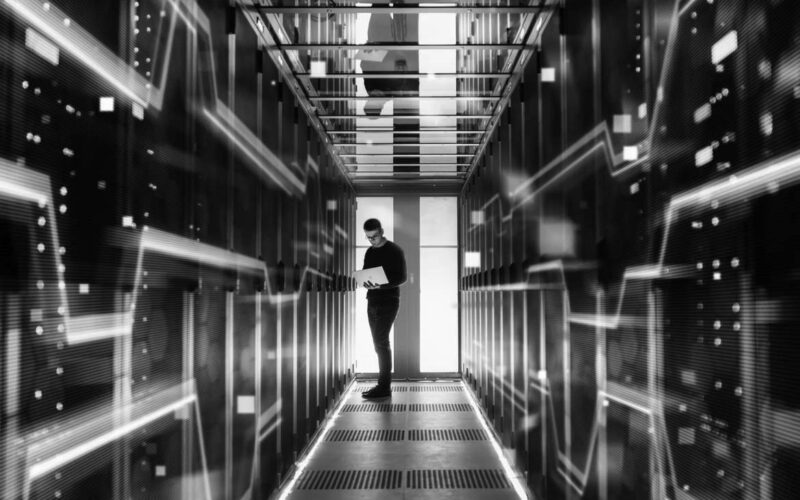 A person stands in a futuristic data center corridor surrounded by glowing blue server racks, using a laptop to create a disaster recovery plan. The scene is illuminated by multiple lights, reflecting a high-tech, digital environment.