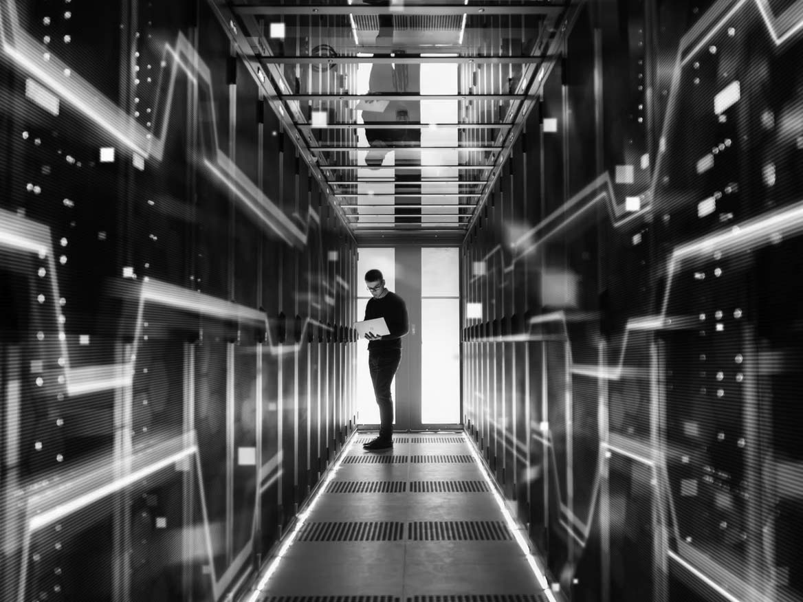 A person stands in a futuristic data center corridor surrounded by glowing blue server racks, using a laptop to create a disaster recovery plan. The scene is illuminated by multiple lights, reflecting a high-tech, digital environment.