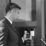 A business professional in a suit holds a smartphone displaying a home security app up to a smart access control panel at a building entrance, showcasing seamless IoT integration while attempting to unlock the door.