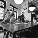 Six people sit around a conference table, listening to a man presenting a proactive approach to cybersecurity at a whiteboard in a modern, well-lit office with large windows, plants, and wooden accents.