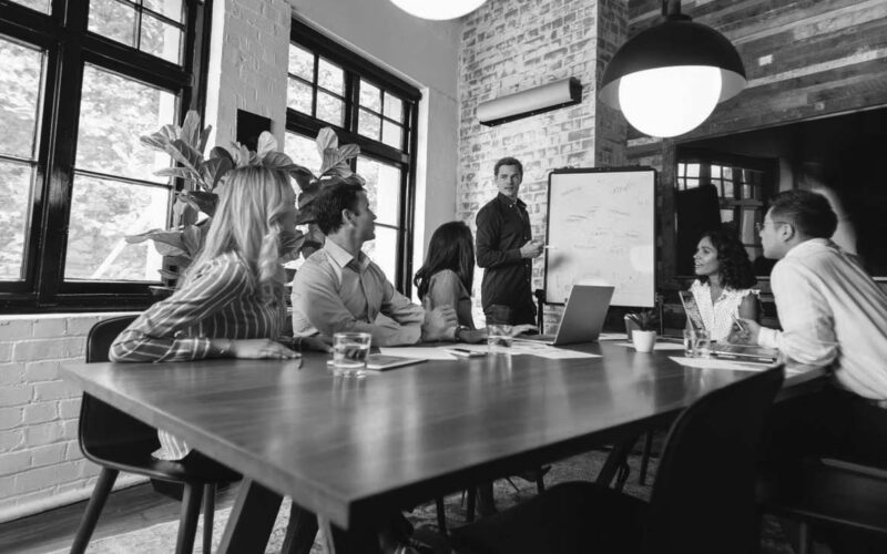 Six people sit around a conference table, listening to a man presenting a proactive approach to cybersecurity at a whiteboard in a modern, well-lit office with large windows, plants, and wooden accents.