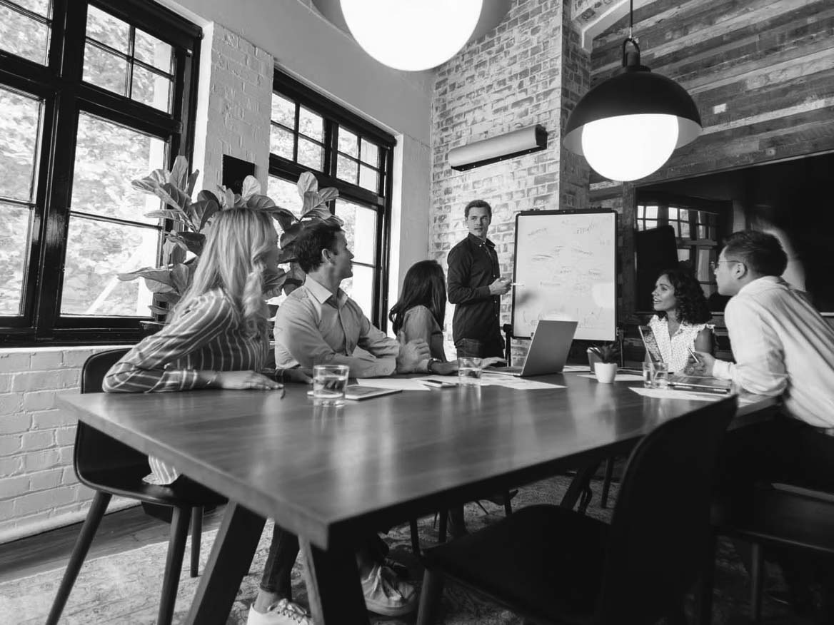 Six people sit around a conference table, listening to a man presenting a proactive approach to cybersecurity at a whiteboard in a modern, well-lit office with large windows, plants, and wooden accents.