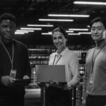 Three people stand in a high-tech server room, each holding a laptop or tablet. Wearing ID badges and business-casual attire, they smile confidently, embodying cyber security as blue server lights glow in the background.