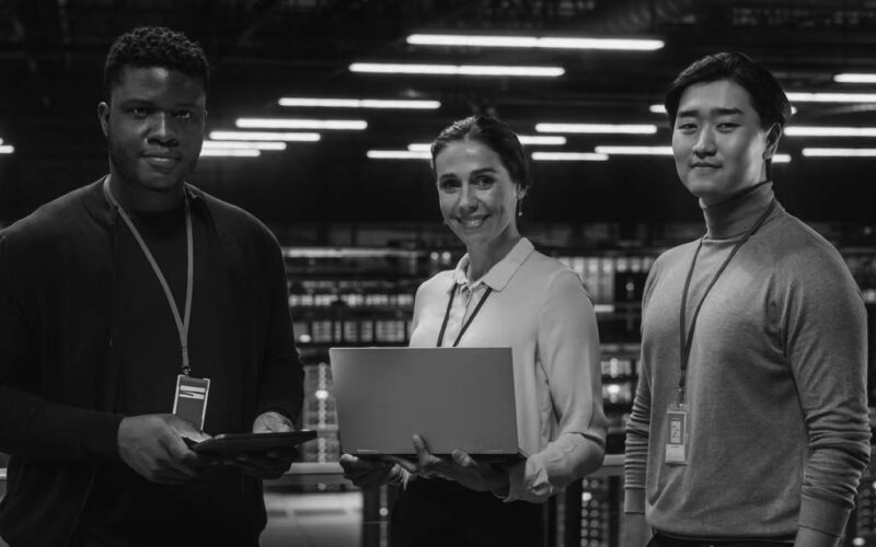 Three people stand in a high-tech server room, each holding a laptop or tablet. Wearing ID badges and business-casual attire, they smile confidently, embodying cyber security as blue server lights glow in the background.